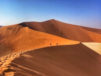 Two People Walking Up A Sand Dune In The Desert